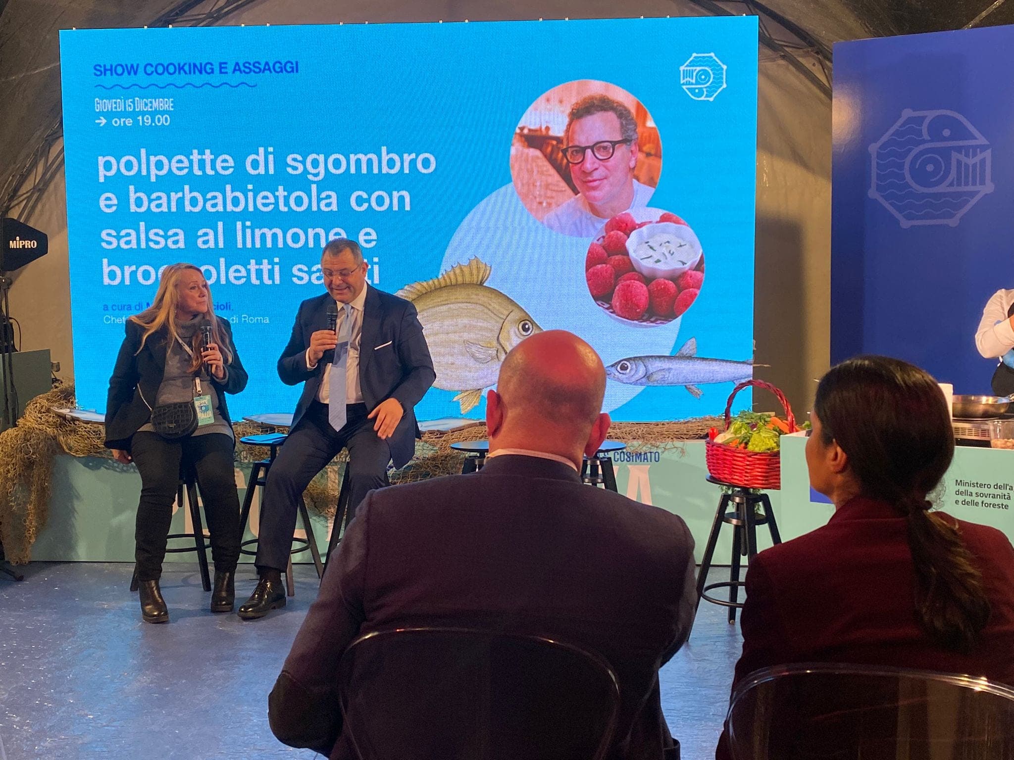 Il palco del convegno con due relatori e lo schermo dello show cooking